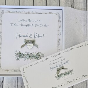 May include: A white card with a green floral border and the text "Wedding Day Wishes To Our Daughter & Son In Law Hannah & Robert" and a silver heart charm with the text "Mr & Mrs". The card is in a white box with a green floral border.