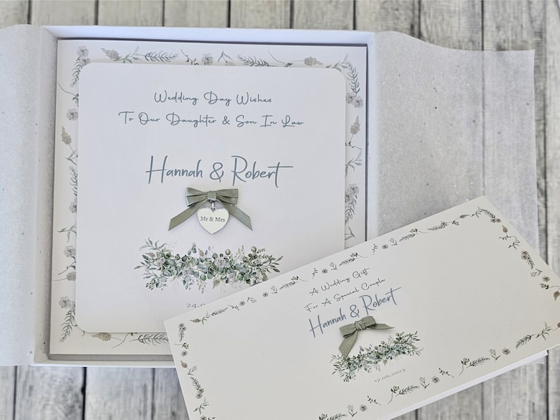 May include: A white card with a green floral border and the text "Wedding Day Wishes To Our Daughter & Son In Law Hannah & Robert" and a silver heart charm with the text "Mr & Mrs". The card is in a white box with a green floral border.