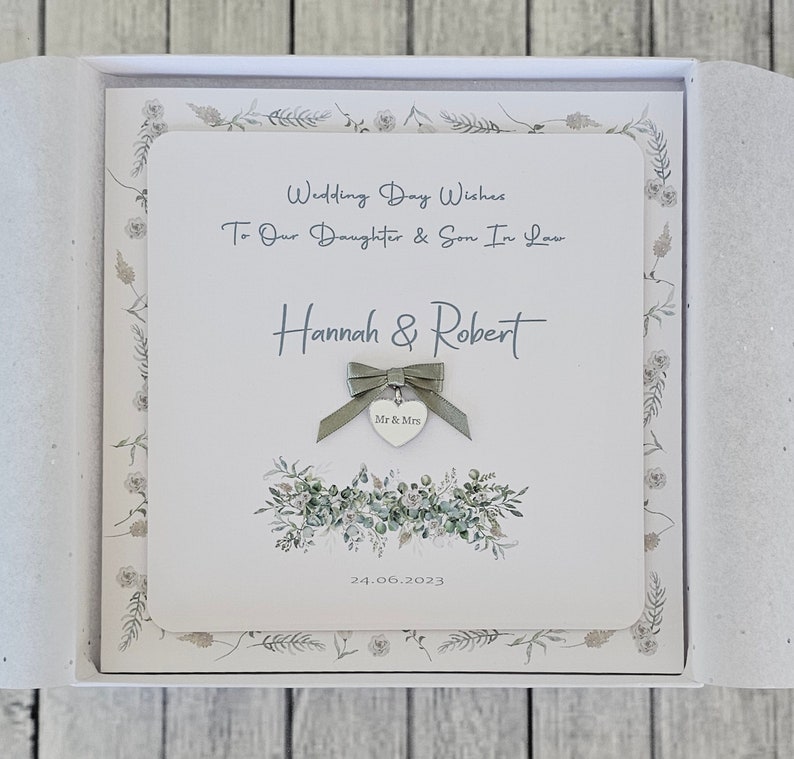May include: A white card with a green floral design and the text "Wedding Day Wishes To Our Daughter & Son In Law Hannah & Robert Mr & Mrs 24.06.2023"