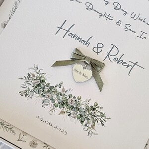 May include: A white card with green foliage and a silver heart charm with the words "Mr & Mrs" on it. The card has the text "To Our Daughter & Son In Wedding Day Wishes Hannah & Robert 24.06.2023" written on it.