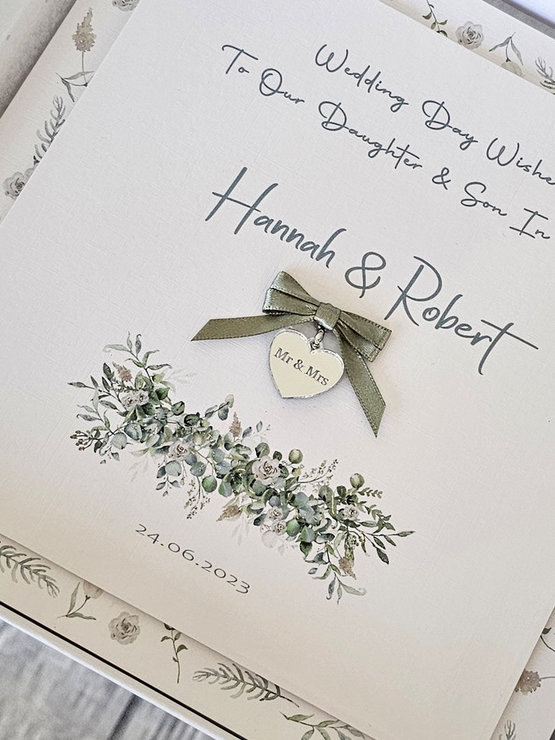 May include: A white card with green foliage and a silver heart charm with the words "Mr & Mrs" on it. The card has the text "To Our Daughter & Son In Wedding Day Wishes Hannah & Robert 24.06.2023" written on it.
