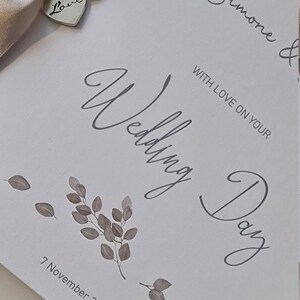 May include: A white card with the text "To our special Simone & Wedding Day" in grey script. The card is decorated with a beige satin ribbon and two silver charms, one with the word "Love" and the other with a heart design. The date "7 November 2021" is printed in grey at the bottom of the card.