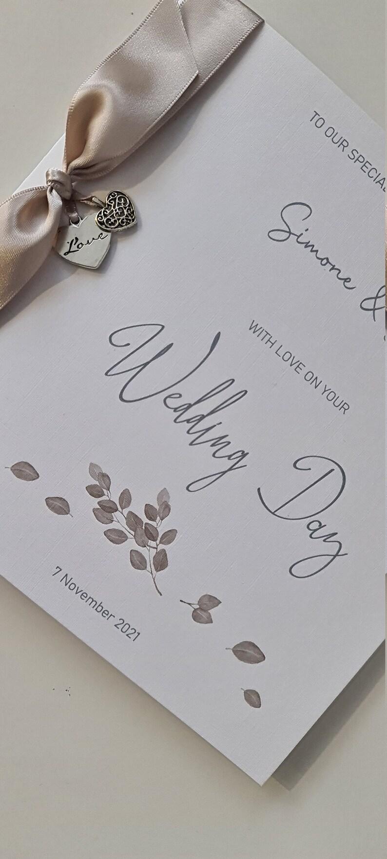 May include: A white card with the text "To our special Simone & Wedding Day" in grey script. The card is decorated with a beige satin ribbon and two silver charms, one with the word "Love" and the other with a heart design. The date "7 November 2021" is printed in grey at the bottom of the card.