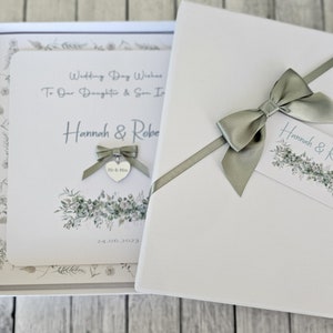 May include: A white card box with a green ribbon and bow. The card has a watercolor design of greenery and the text "Wedding Day Wishes To Our Daughter & Son In Law Hannah & Robert 24.06.2023".