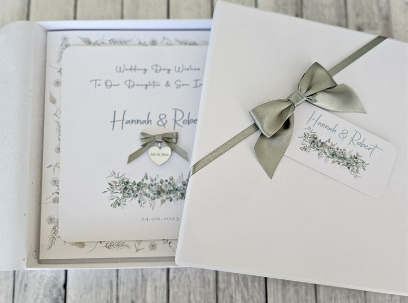 May include: A white card box with a green ribbon and bow. The card has a watercolor design of greenery and the text "Wedding Day Wishes To Our Daughter & Son In Law Hannah & Robert 24.06.2023".