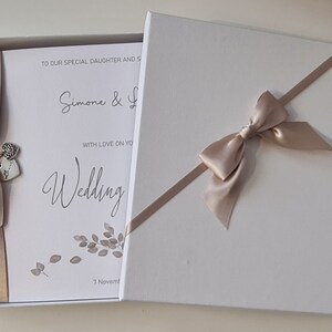 May include: A white box with a beige ribbon and bow. The box contains a white card with the text "To our special daughter and son, Simone & [unclear text], with love on your wedding, 7 November".