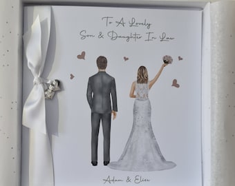 Puede incluir: Una tarjeta blanca con una cinta plateada y un dije que dice "Bride & Groom". La tarjeta tiene una ilustración de acuarela de una novia y un novio con su atuendo de boda. El texto en la tarjeta dice "To A Lovely Son & Daughter In Law" y "Adam & Elise".