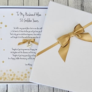 Pode incluir: Um cartão branco com confete dourado e uma fita dourada amarrada ao redor. O cartão diz "To My Husband Alan 50 Golden Years".