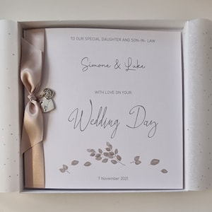 May include: A white card with a beige ribbon and silver heart charm. The card reads "To our special daughter and son-in-law Simone & Luke With love on your Wedding Day 7 November 2021".
