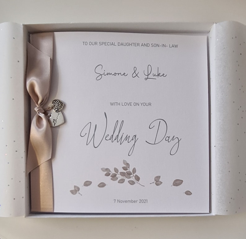 May include: A white card with a beige ribbon and silver heart charm. The card reads "To our special daughter and son-in-law Simone & Luke With love on your Wedding Day 7 November 2021".