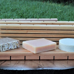 May include: Three bars of soap on a wooden soap dish. The soap dish has slats to allow water to drain. The soap bars are different colors and shapes. The soap dish is on a wooden surface.