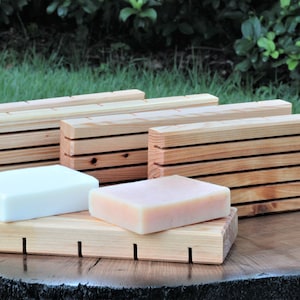 May include: Two bars of soap, one white and one light pink, are resting on a wooden soap dish. The dish has a series of slots to help the soap drain. Behind the dish are four wooden soap savers, stacked in a row.