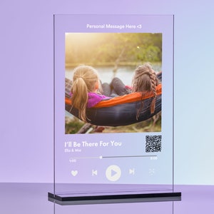 以下が含まれることがあります： ハンモックに乗った2人の女の子の写真が描かれた透明なガラスのプレート。プレートには、Ella & Miaの曲「I'll Be There For You」が再生されている音楽プレーヤーのインターフェースが表示されています。プレートにはQRコードも含まれています。