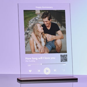 May include: A clear glass photo frame with a couple in a photo and a music player interface with the song "How long will I love you" by Ellie Goulding playing. The frame has a wooden base.