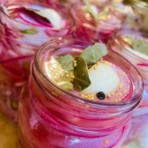 May include: Close-up of several glass jars filled with pickled red onions. The onions are a vibrant pink color and are submerged in a clear brine. Spices and herbs, including bay leaves and mustard seeds, are visible.