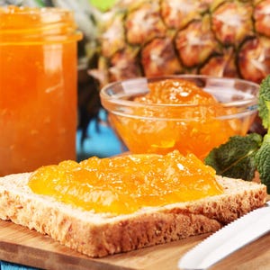 May include: A slice of bread topped with a generous amount of orange marmalade. The bread is on a wooden cutting board with a knife nearby. There is a jar of marmalade and a pineapple in the background.