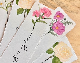 May include: Five watercolor paintings of different flowers on white paper with torn edges. The flowers are a white daisy, a yellow rose, a pink carnation, a purple cosmos, and a yellow rose. The paintings are arranged in a fan shape with the names "Suzie" and "Sin" written in black ink on the bottom of the paintings.