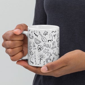 May include: White ceramic mug with a black and white doodle design featuring various symbols, including a musical instrument, a boot, a hat, and the words "Qazaq" and "Salem".