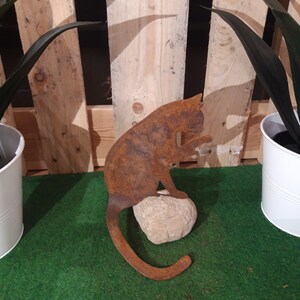 Könnte beinhalten: Eine rostige Metallkatzenskulptur, die auf einem Stein sitzt. Die Katze ist in einer zusammengerollten Position mit dem Schwanz um den Körper gewickelt. Die Skulptur befindet sich auf einer grünen künstlichen Grasfläche.