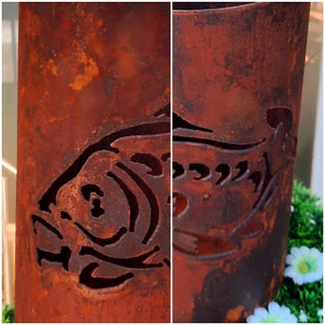Könnte beinhalten: Ein rostiger Metalltopf mit einem ausgeschnittenen Fischdesign. Der Topf ist zylindrisch und hat eine dunkelbraune Rostfarbe.