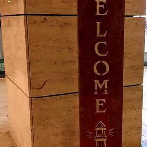 Könnte beinhalten: Ein rostiges Metallschild mit dem Wort "WELCOME" in weißen Buchstaben. Das Schild zeigt auch eine weiße Silhouette eines Leuchtturms mit Vögeln, die um ihn herum fliegen.