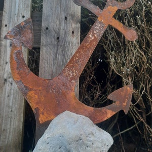 Könnte beinhalten: Ein rostiger Anker aus Metall mit abgerundeter Oberseite und spitzer Unterseite, der auf einem grauen Stein ruht. Der Anker ist ein beliebtes nautisches Dekorationselement.