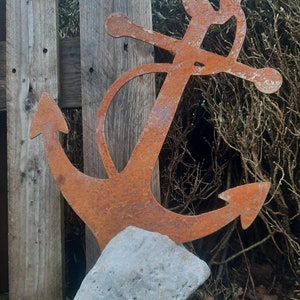 Könnte beinhalten: Eine rostige Metallanker-Skulptur mit einem verwitterten grauen Steinsockel. Der Anker ist traditionell gestaltet mit einem Querstück und einem Ring oben.