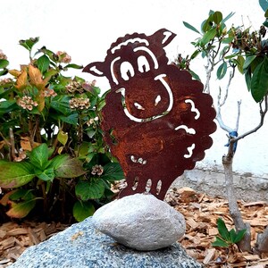 Könnte beinhalten: Eine rostige Metall-Schafskulptur mit einem lächelnden Gesicht, die auf einem grauen Stein steht. Die Skulptur befindet sich in einem Garten mit grünen Pflanzen und braunen Holzspänen.