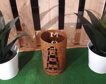 May include: A rusty metal planter with a lighthouse design and the letter "M" cut out. The planter is sitting on a green surface.