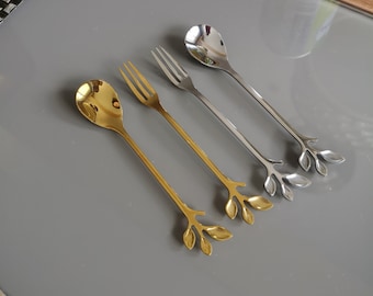 May include: Set of four gold and silver leaf-shaped spoons. The spoons have a long, thin handle with a small, round bowl at the end. The handles are decorated with a leaf design.