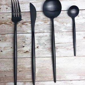 Pode incluir: Um conjunto de quatro peças de talheres pretos, incluindo um garfo, uma faca, uma colher de sopa e uma colher de chá, dispostos sobre uma superfície de madeira clara.