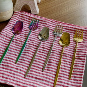 May include: Five iridescent stainless steel utensils with rose-shaped handles on a red and white striped cloth. The utensils include a spoon, a fork, and three spoons.