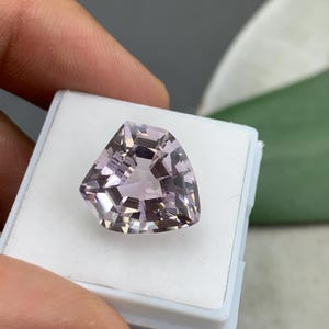 以下が含まれることがあります： ユニークな三角形の形をした、ファセット加工された薄紫色の宝石。 宝石は白いプラスチックケースに入れられており、その複雑なカットと反射面が示されています。 宝石の色は柔らかく淡い紫色です。