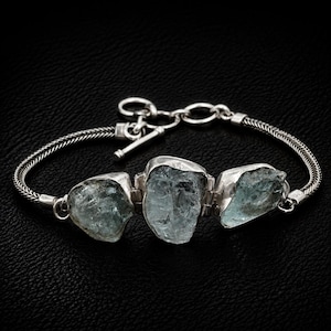 May include: A silver bracelet featuring three raw, light blue aquamarine stones set in metal bezels. The bracelet is constructed with a woven chain and includes a toggle clasp closure with a circular ring and a T-bar component for fastening.