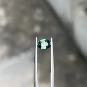 May include: An oval-cut gemstone displaying a gradient of deep teal and vibrant green hues. The faceted stone is held securely between the serrated metal tips of a pair of silver-toned tweezers, showcasing its clarity and reflective surface.