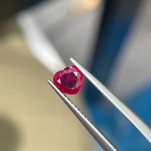 Könnte beinhalten: Ein herzförmiger, facettierter Rubin, der von einer Pinzette gehalten wird. Der Rubin hat eine tiefrote Farbe und eine polierte Oberfläche. Die Pinzette ist silberfarben und hält den Edelstein vor einem unscharfen Hintergrund.