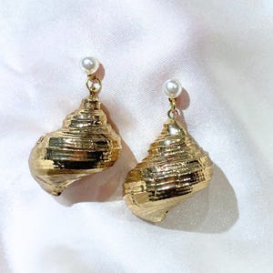 Può includere: Un paio di orecchini a forma di conchiglia dorata con un piccolo accento di perla sulla parte superiore. Gli orecchini hanno una superficie strutturata e sono esposti su uno sfondo bianco. Gioielli.