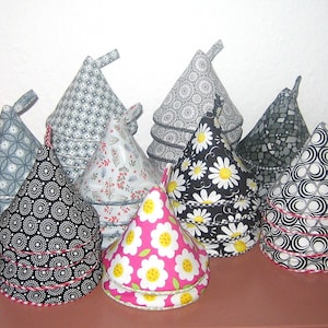 Könnte beinhalten: Eine Sammlung von kegelförmigen Teewärmern in verschiedenen Mustern und Farben. Designs umfassen Blumen-, geometrische und Gänseblümchen-Prints. Die Wärmer sind aus Stoff und haben eine Schlaufe oben. Sie sind auf einer braunen Oberfläche angeordnet.
