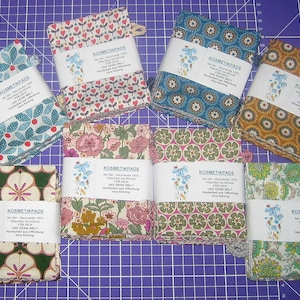 Könnte beinhalten: Ein Set mit acht handgefertigten wiederverwendbaren Kosmetikpads aus Baumwolle in verschiedenen floralen und geometrischen Mustern. Die Pads sind in Stapeln zu vieren verpackt und mit "Kosmetikpads" beschriftet, mit dem Text "Ein Set - Baumwolle 100%  Waschbar für dich und deine Welt  Handarbeit aus Oldenburg  Vera Kunnig".