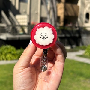 May include: A red and white retractable badge reel featuring a cartoon dog face. The reel has a clear plastic strap and a black cord. The dog face is white with black details and is set against a red background.