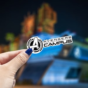 May include: A white sticker with a black Avengers Campus logo. The logo features a stylized "A" with an arrow pointing to the right, surrounded by a circle. The text "Avengers Campus" is written in black letters below the "A".