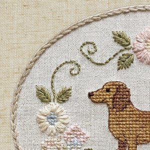 May include: Embroidered hoop art featuring a brown dachshund dog, surrounded by embroidered flowers and green foliage. The design is set against a white fabric background, with a braided border.