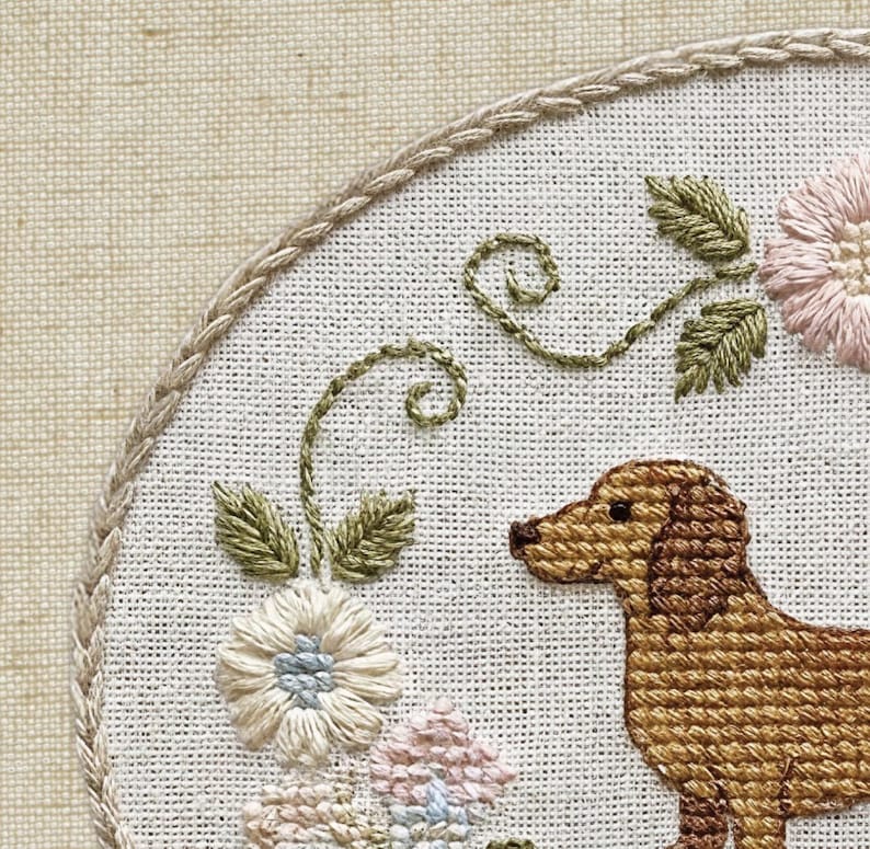 May include: Embroidered hoop art featuring a brown dachshund dog, surrounded by embroidered flowers and green foliage. The design is set against a white fabric background, with a braided border.