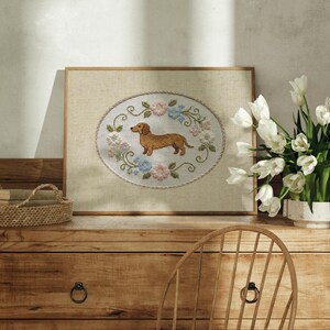 May include: A framed cross-stitch artwork featuring a dachshund dog in the center, surrounded by a floral wreath of pink, blue, and green. The artwork is displayed on a wooden dresser, with a basket and a vase of white tulips nearby.