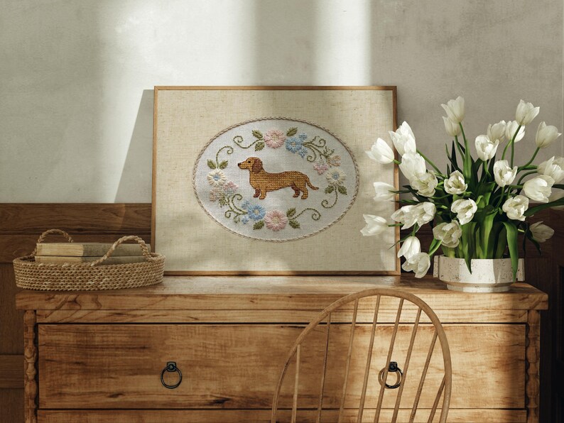 May include: A framed cross-stitch artwork featuring a dachshund dog in the center, surrounded by a floral wreath of pink, blue, and green. The artwork is displayed on a wooden dresser, with a basket and a vase of white tulips nearby.