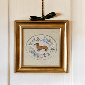 May include: A framed, embroidered artwork featuring a brown dachshund dog in the center, surrounded by a floral wreath of pink and blue flowers. The artwork is set in a gold-colored frame and hangs from a green ribbon.