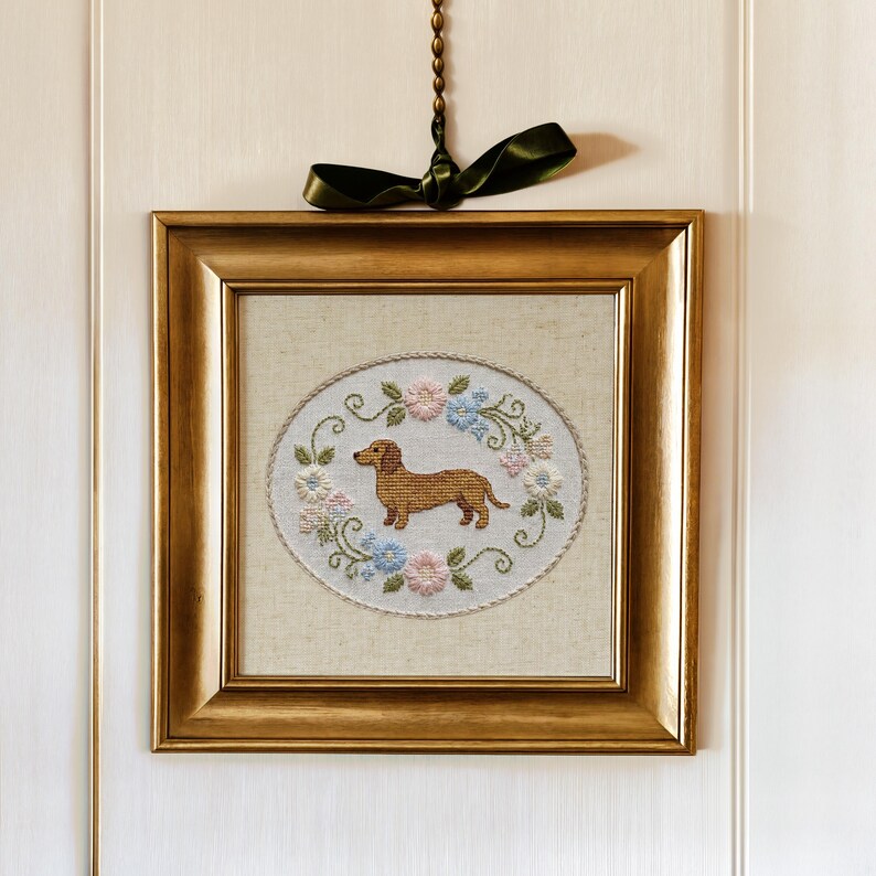 May include: A framed, embroidered artwork featuring a brown dachshund dog in the center, surrounded by a floral wreath of pink and blue flowers. The artwork is set in a gold-colored frame and hangs from a green ribbon.