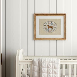 May include: A framed embroidery featuring a brown dachshund dog within an oval design, surrounded by floral accents in a light wood frame. The artwork hangs above a white crib with a quilted blanket.