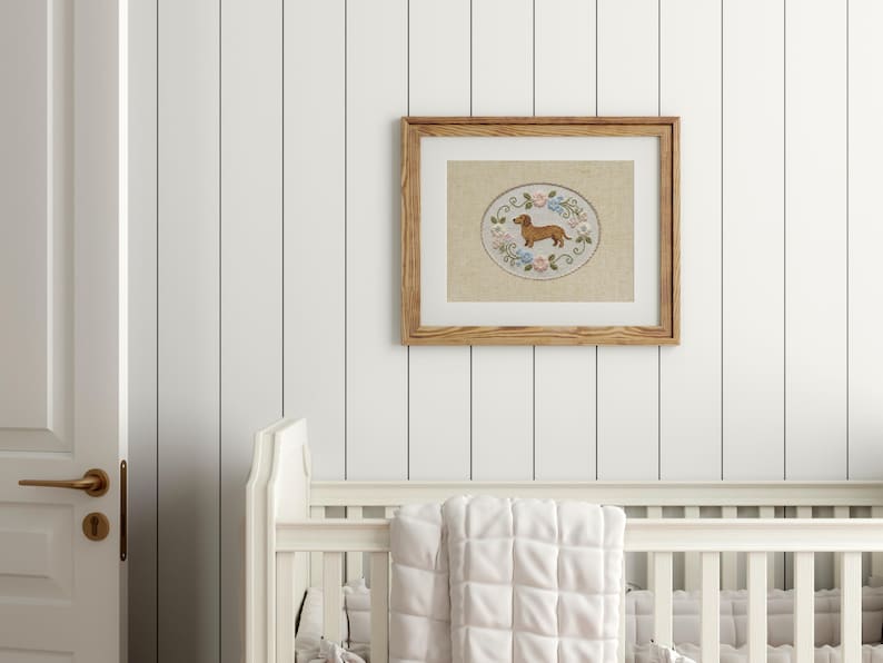 May include: A framed embroidery featuring a brown dachshund dog within an oval design, surrounded by floral accents in a light wood frame. The artwork hangs above a white crib with a quilted blanket.