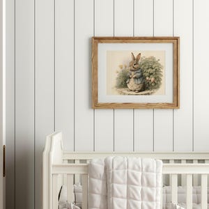 Peut inclure: Une illustration encadr&eacute;e d'un lapin brun portant une veste bleue et tenant une fleur. Le lapin se tient dans un jardin avec des feuilles vertes et des fleurs roses. Le cadre est en bois et a une finition brun clair.